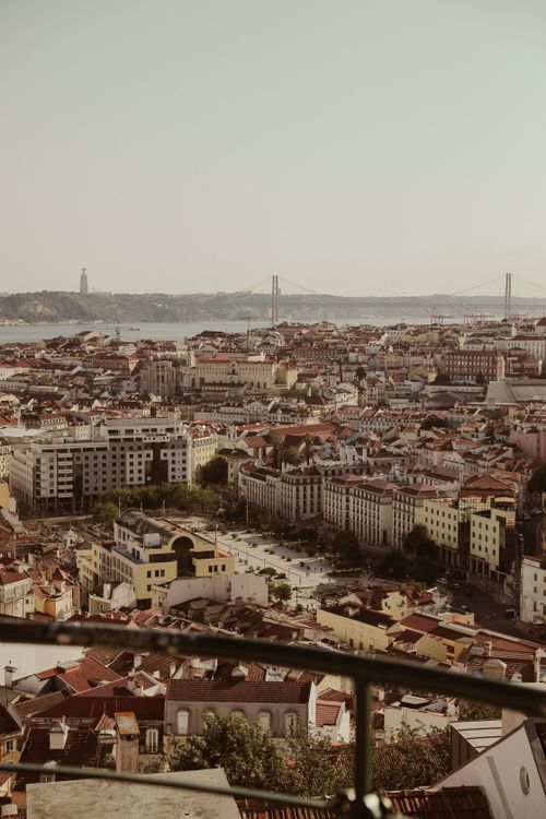 Vista panorâmica de Lisboa com a Ponte 25 de Abril e o Rio Tejo ao fundo, com os telhados típicos da cidade em primeiro plano