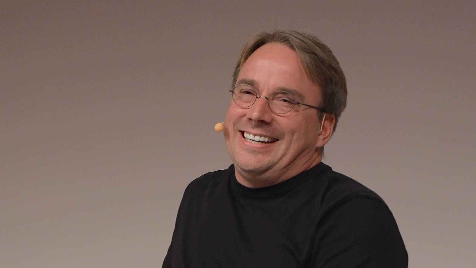 Retrato de Linus Torvalds, de óculos e camisola preta, a rir com um microfone de lapela, diante de um fundo claro e neutro.