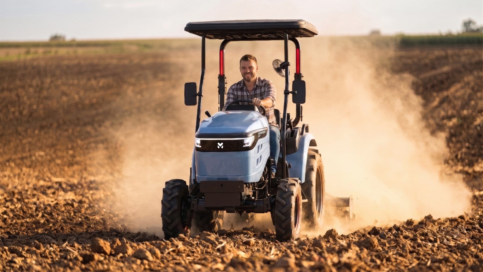 Pessoa conduz um tractor compacto azul por um campo de solo revolvido