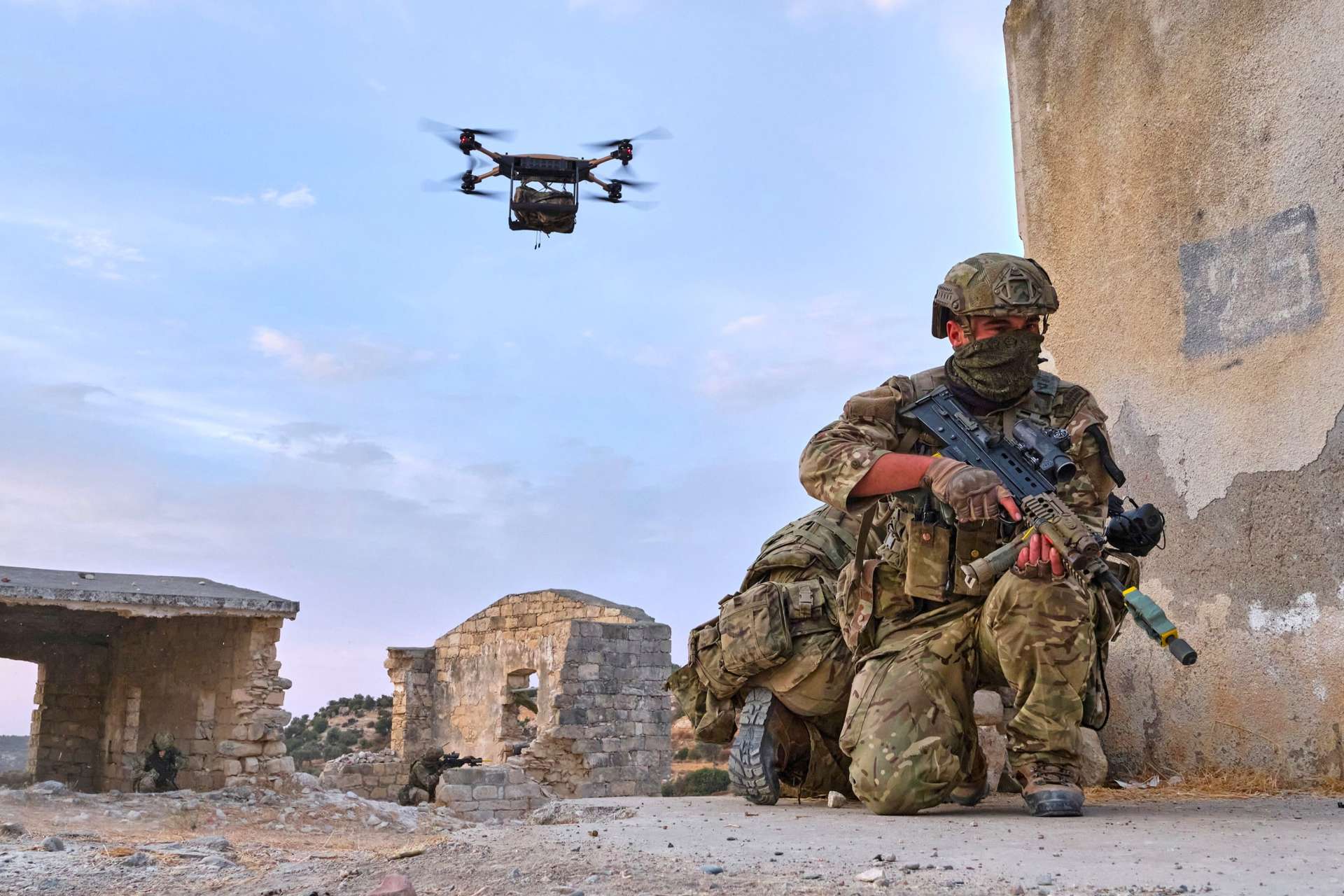 Soldado britânico em cenário de conflito com um drone militar quadcopter sobrevoando — a nova era de guerra autónoma que o investimento de £5 mil milhões do Reino Unido pretende acelerar