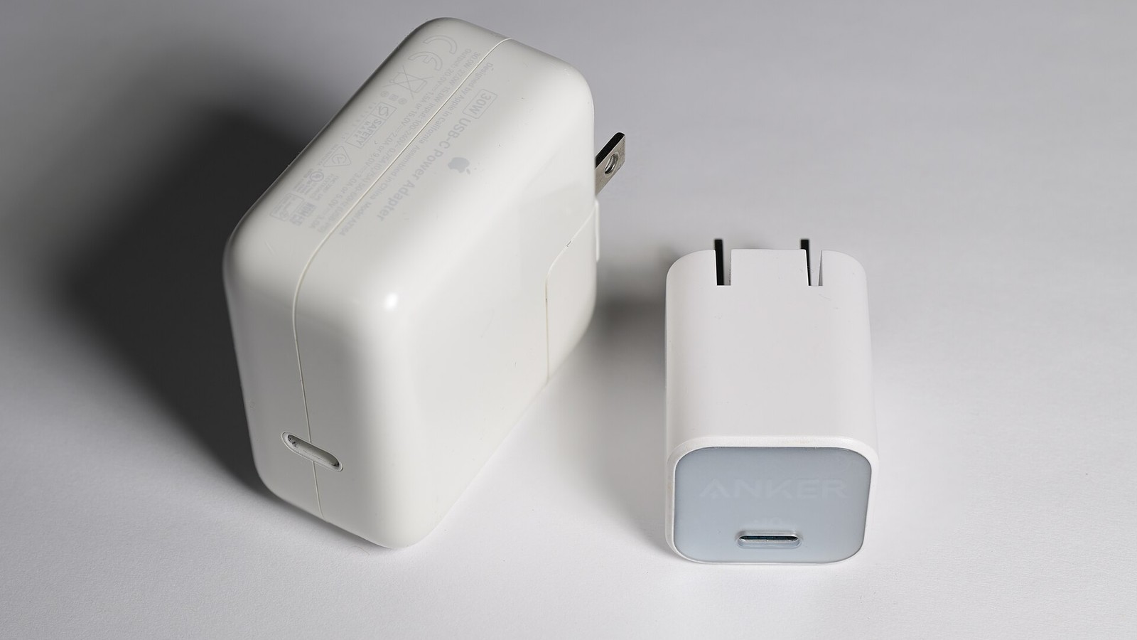 Dois carregadores USB-C de 30 W sobre fundo claro: à esquerda um carregador Apple, mais alto e volumoso, à direita um Anker com marca GaN, quase metade do volume.