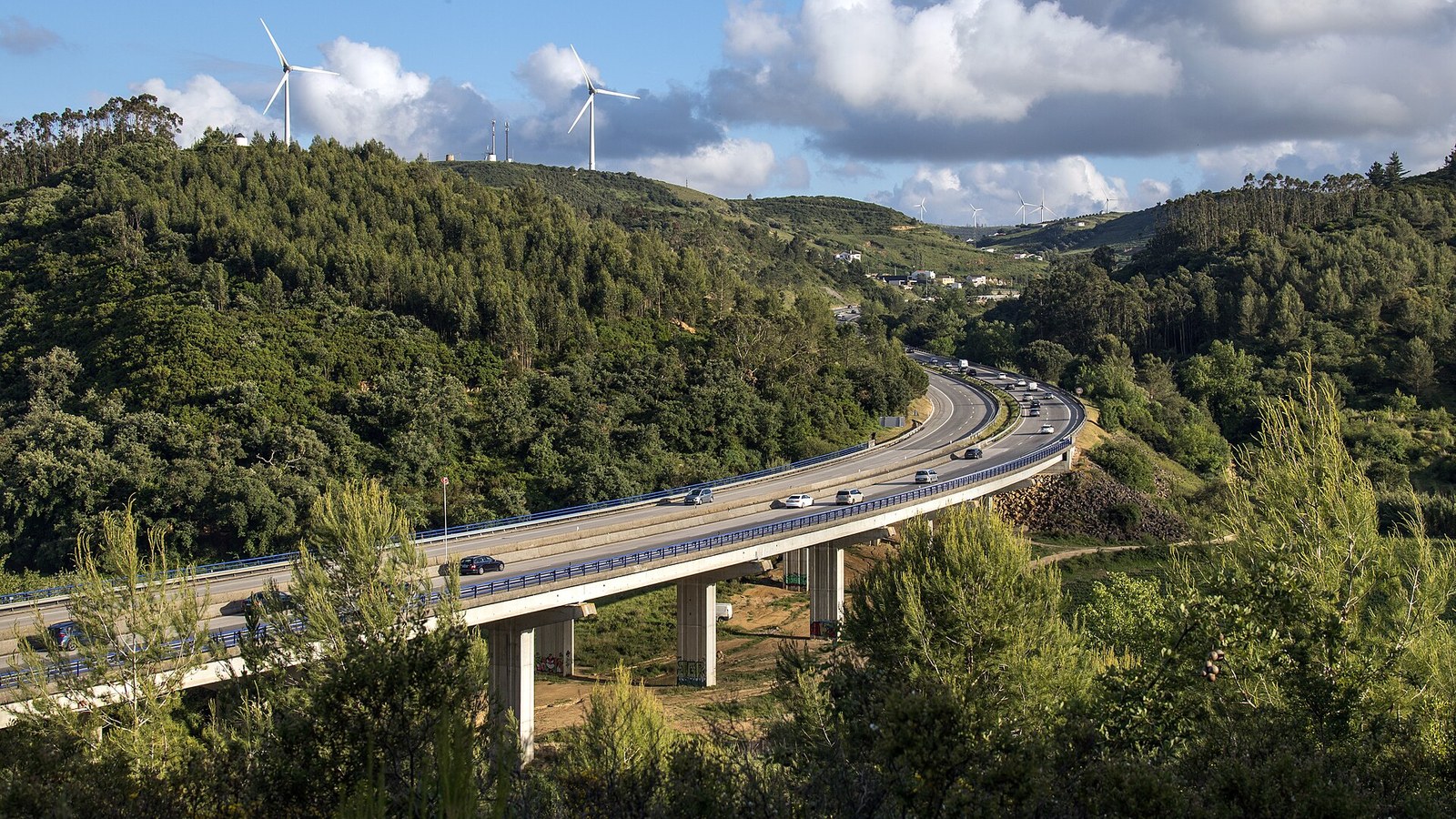 Autoestrada A8 em viaduto sobre um vale arborizado, com trânsito nos dois sentidos e turbinas eólicas nas colinas ao fundo