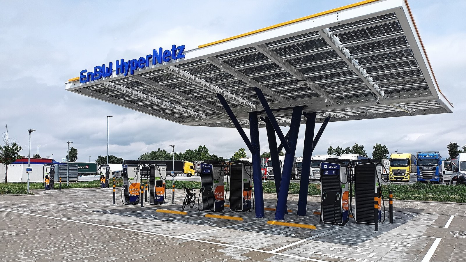 Parque de carregamento ultrarrápido com cobertura de painéis solares sobre várias colunas de carregamento de 400 kW e camiões parados ao fundo